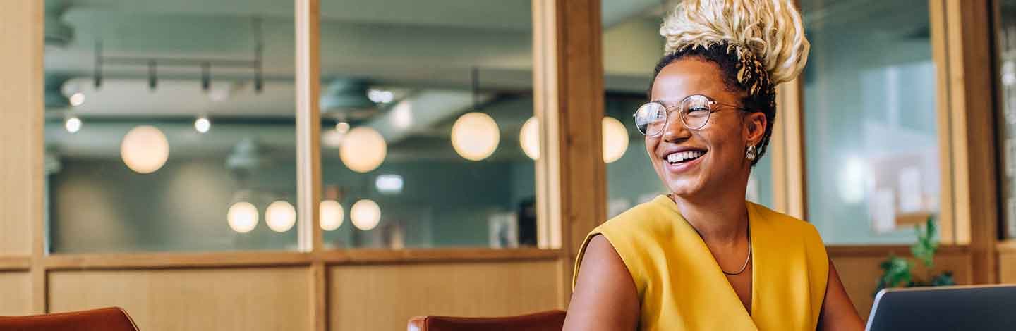 Smiling woman with blonde updo hairstyle wearing glasses and yellow sleeveless top in modern office with glass walls.