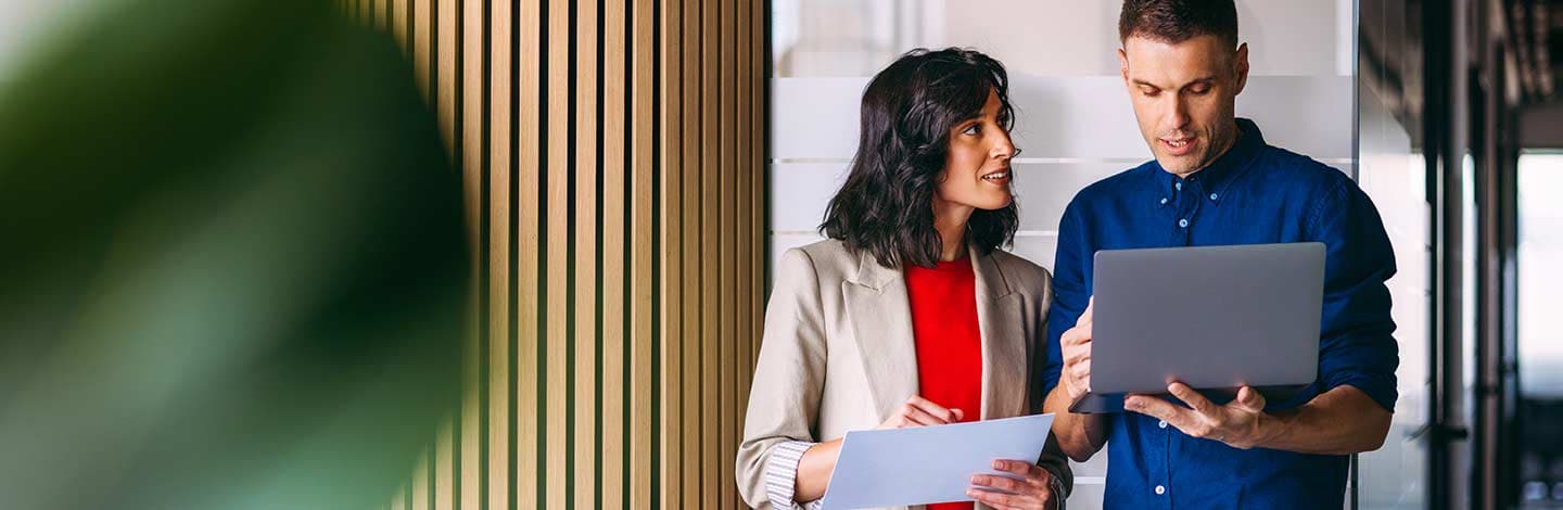 Two business professionals collaborating with laptop and documents in modern office with wood paneling and white brick wall.