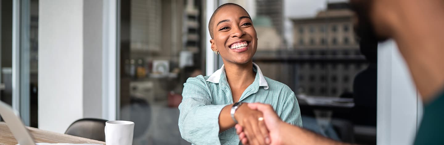 Smiling woman with short hair in blue striped shirt shaking hands across a table in a modern office setting.
