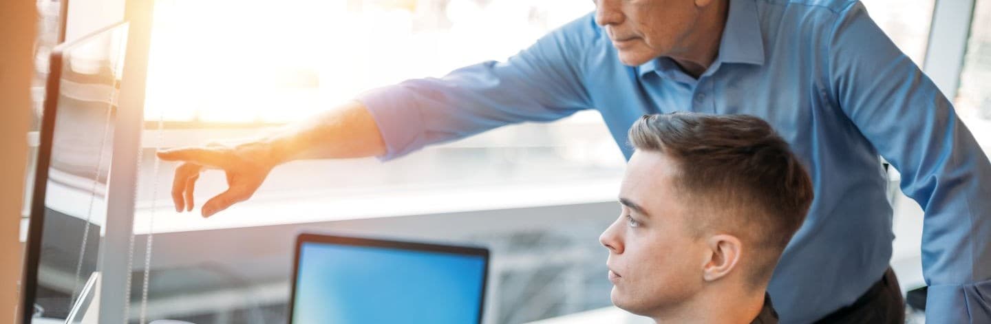 An image of an older man in a blue shirt directing a younger man in a checked shirt on how to run a programme on a desktop computer.