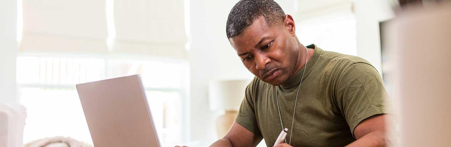 A person sits at a table writing on paper with one hand while using a laptop with the other, in a bright room with natural light.
