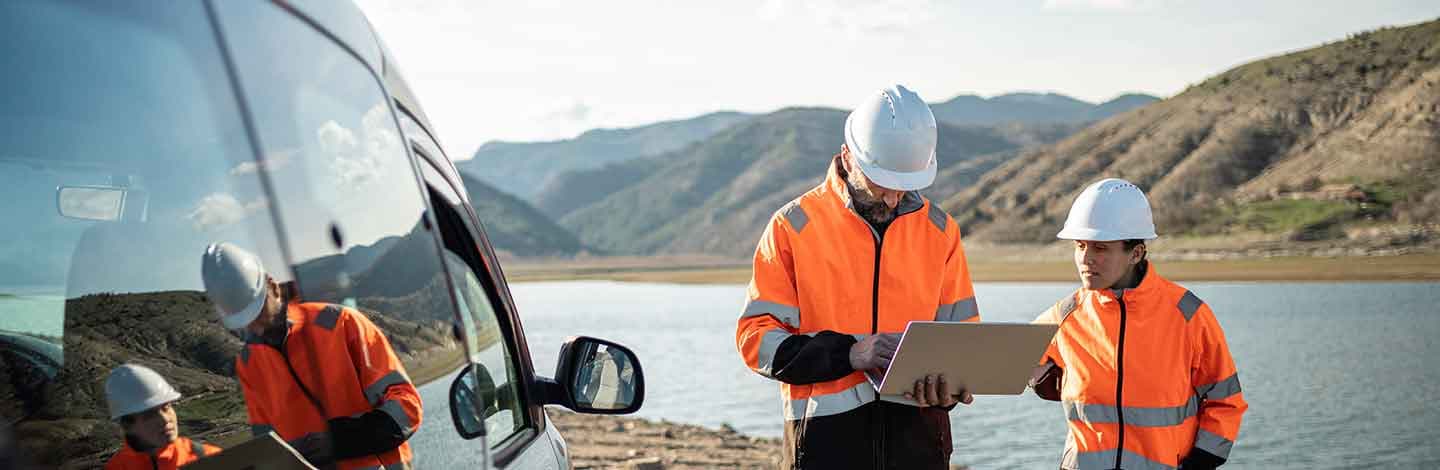 Two workers in orange safety vests and white hard hats review a laptop near a vehicle by a lake with mountains behind.