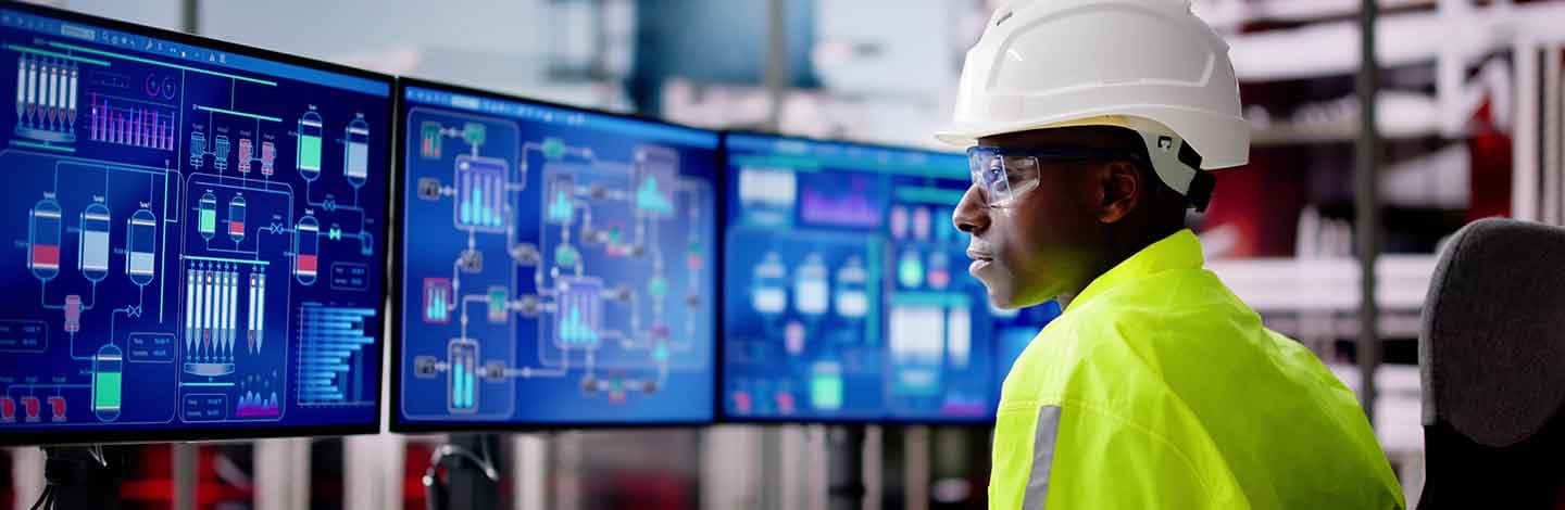 Industrial worker in white hard hat and yellow safety vest monitoring multiple blue computer screens displaying process data.