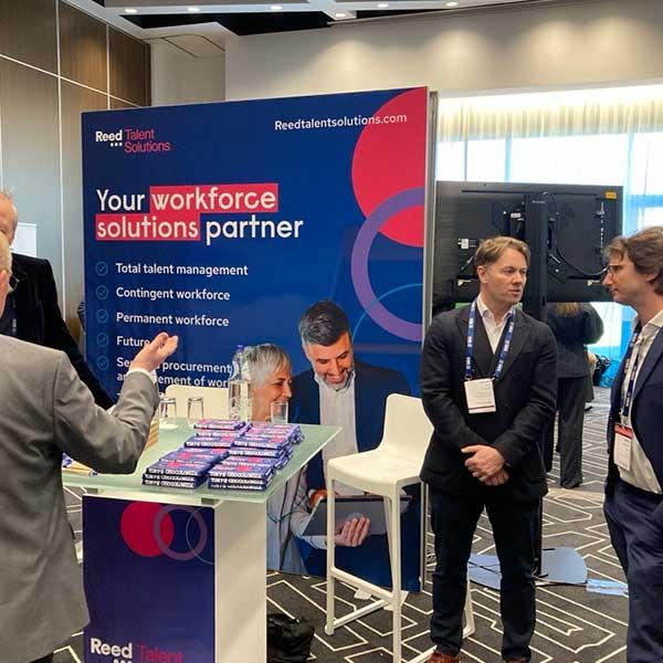Reed Talent Solutions booth at trade show with blue banner displaying workforce solutions services and attendees networking.