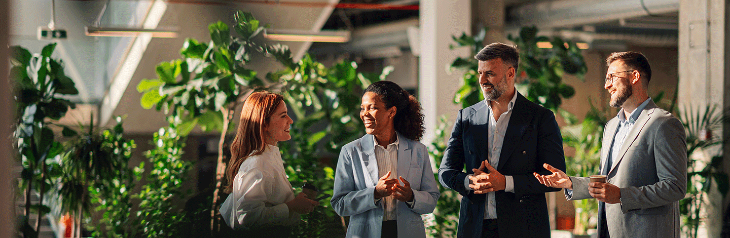 Four business professionals in suits having a friendly conversation in a modern office with abundant green plants.