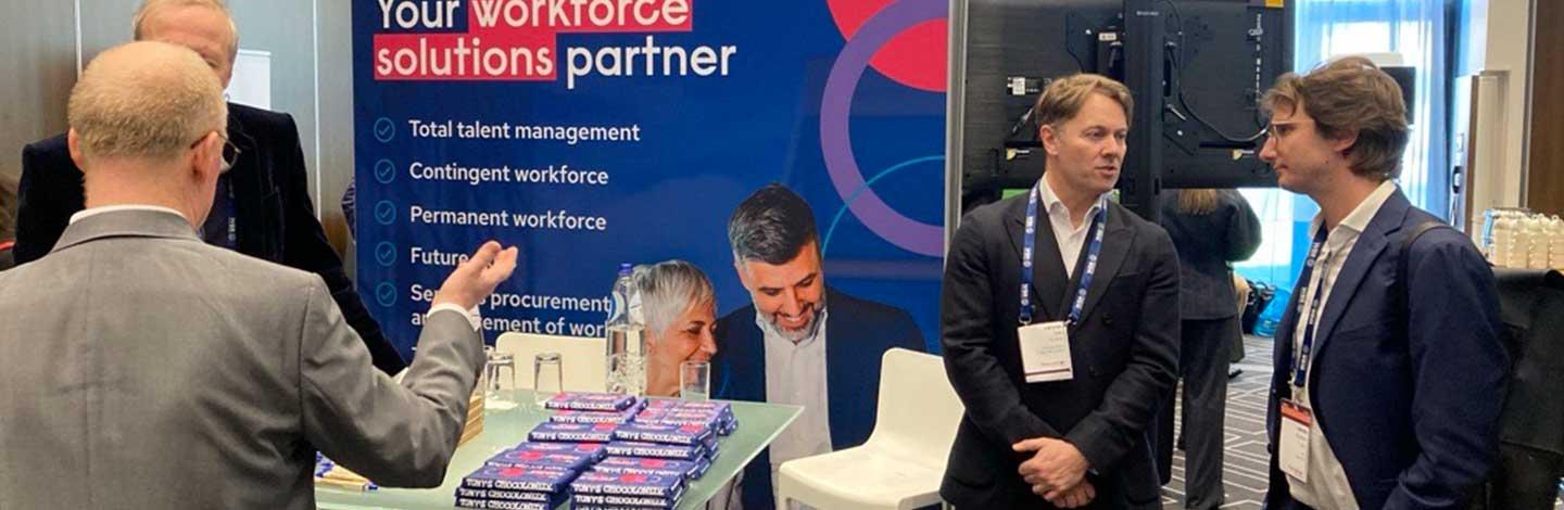 Business professionals networking at a trade show booth with blue workforce solutions banner and promotional materials.