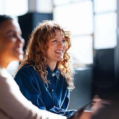 Woman with curly hair in blue shirt smiling during office meeting with colleagues in bright workspace