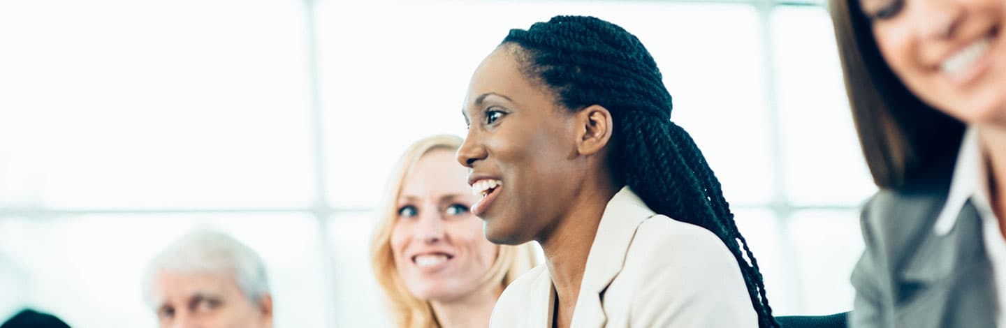 Smiling woman with braided hair in beige blazer during a business meeting with colleagues in bright office setting.