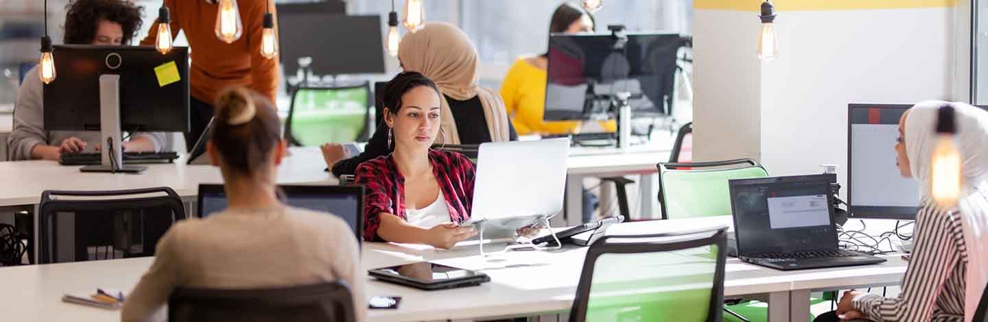Diverse team working at computers in modern open office with pendant lighting and colorful workstations.
