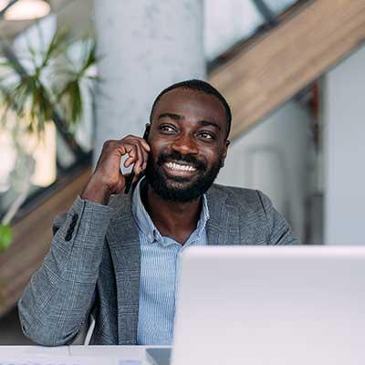 A person sits at a desk in a modern office, speaking on a mobile phone while working on a laptop. A staircase and indoor plants are visible in the background.
