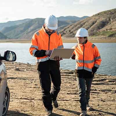 Two workers in orange safety vests and white hard hats review a laptop near a lake with mountains in the background.