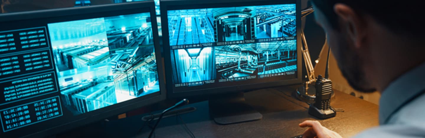 Person monitoring multiple security camera feeds on computer screens showing blue-tinted surveillance footage of corridors.