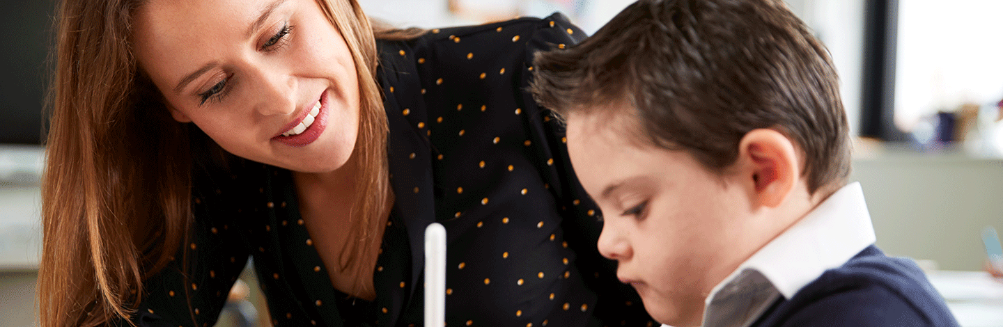 Woman in black polka dot top smiling while working with a young boy in school uniform at a desk.