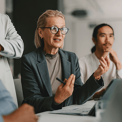 Businesswoman with glasses in gray suit gestures while speaking during a meeting with colleagues in modern office setting.