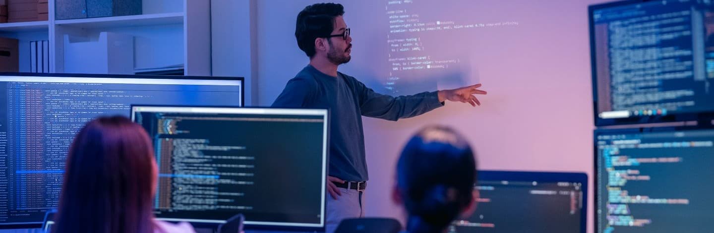 A man wearing glasses in front of a projection of lines of code teaching two people who are also looking at their own laptops.