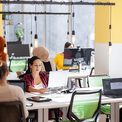 Modern office workspace with people at desks, hanging Edison bulbs, yellow accent wall, and green chairs in foreground.