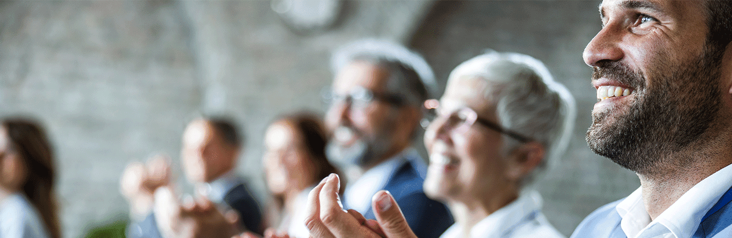 Smiling bearded man in business attire with blurred audience members in background during a presentation or meeting.
