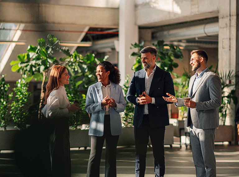 Four business professionals in suits having a conversation in a modern office space with green plants in the background.