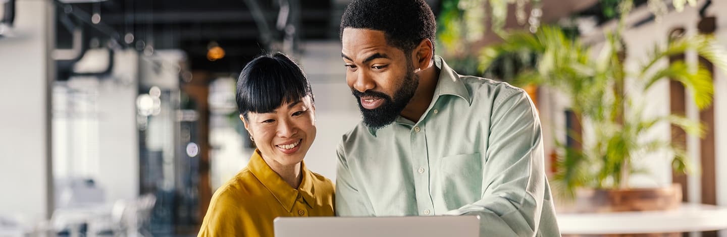 Two smiling professionals, a woman in a yellow shirt and a man in a green shirt, looking at a laptop together in an office.