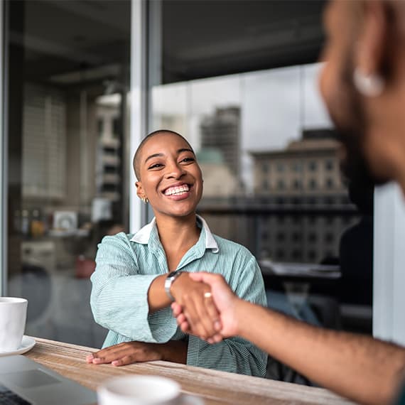 Smiling woman in light blue shirt shaking hands with another person across a table in a modern office setting.