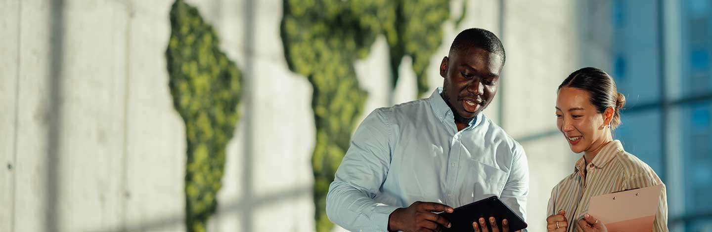 Two colleagues, standing in a modern office setting with a green foliage world map on the wall behind them. The man holds a tablet and gestures while speaking, and the woman smiles, holding a pink folder.