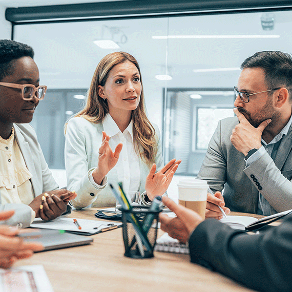 Business professionals in meeting, woman in white shirt gesturing while speaking to colleagues around conference table.