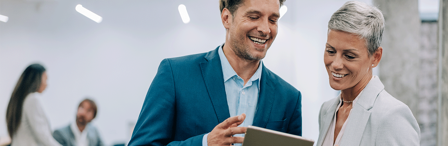 Two smiling business professionals in suits looking at a tablet together in a modern office setting.