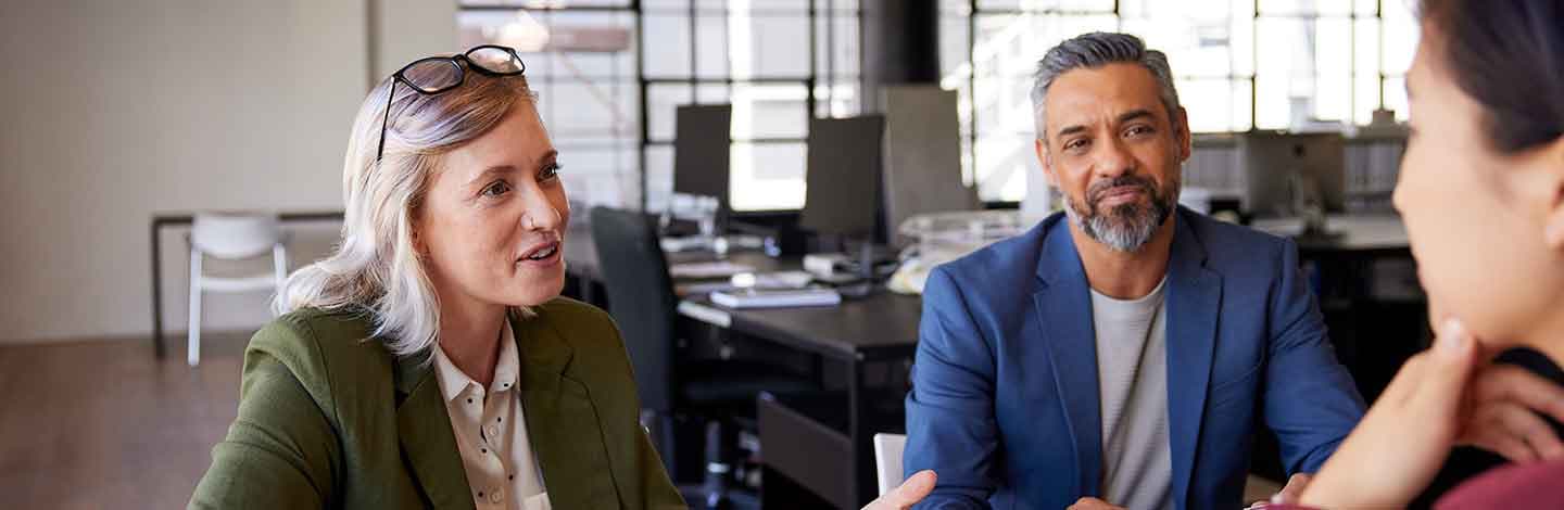 Woman in olive blazer and man in blue blazer engaged in conversation in modern office with glass partitions.