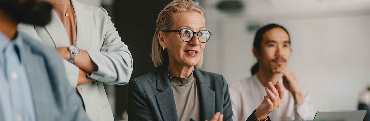 Professional woman with glasses in gray blazer speaking during business meeting with colleagues listening