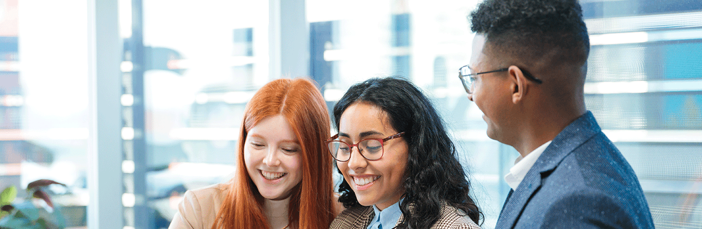 Three diverse professionals smiling while looking at something together in a bright modern office with windows.
