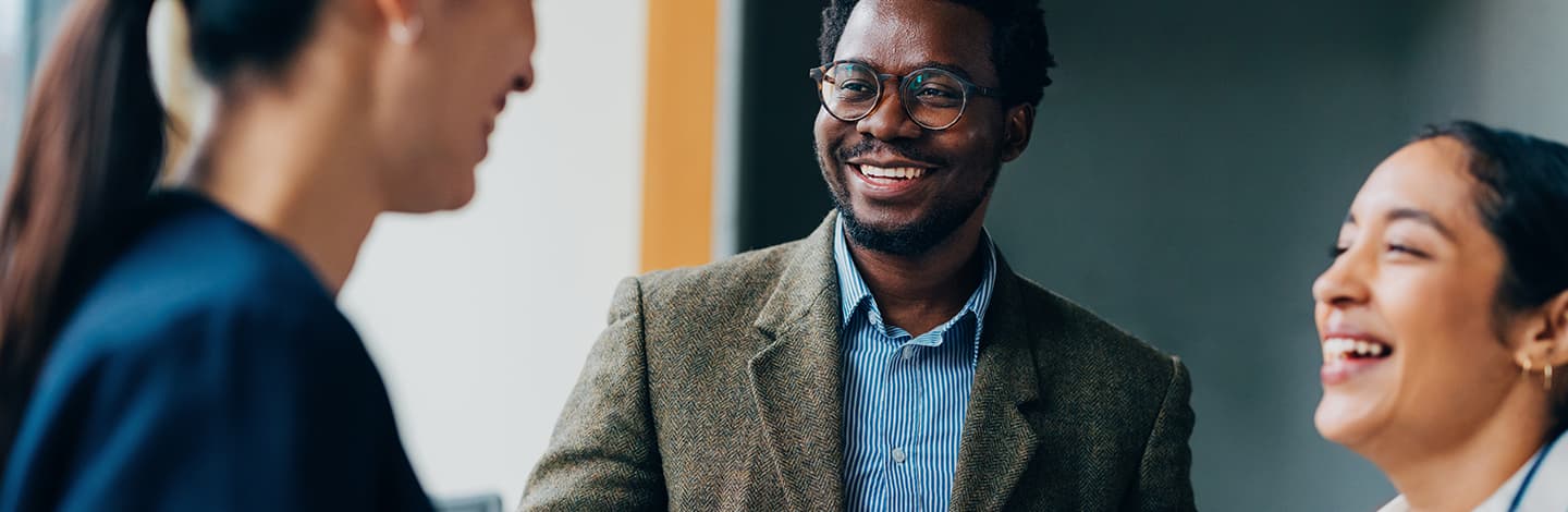 Smiling man in glasses and tweed blazer engaged in conversation with two colleagues in a professional office setting.