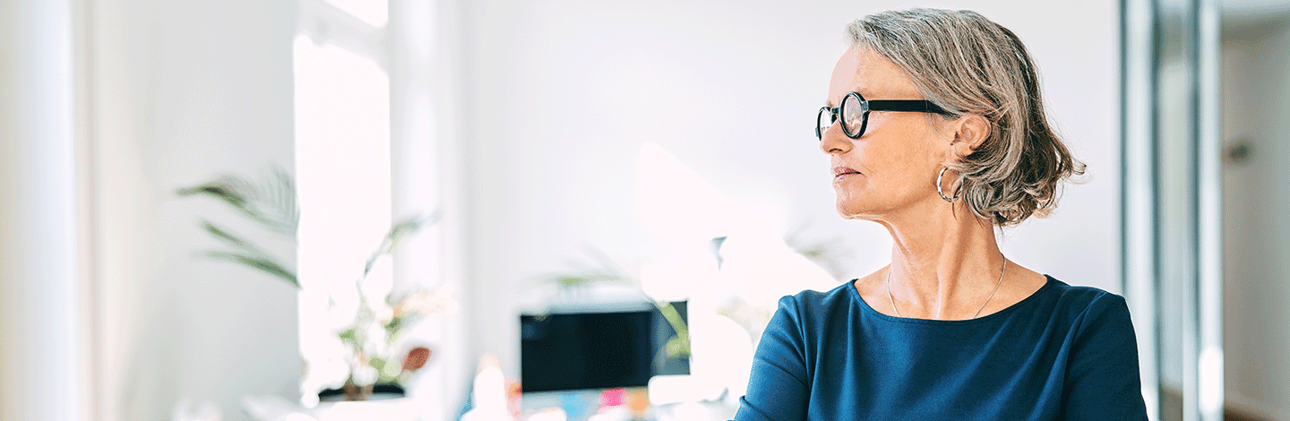 Professional woman with gray hair and round glasses wearing a teal blue top in a bright modern office setting.