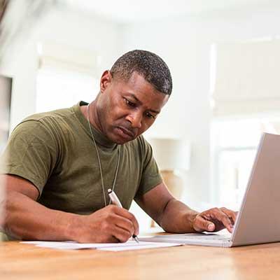 A person sits at a table writing on paper with one hand while using a laptop with the other, in a bright room with natural light.