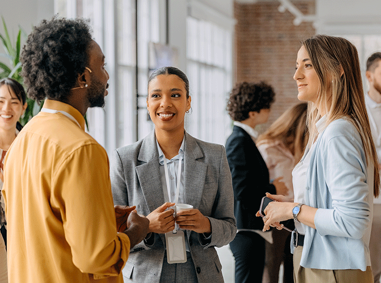 Three diverse professionals networking in a bright modern office, smiling woman in gray suit holding coffee at center.