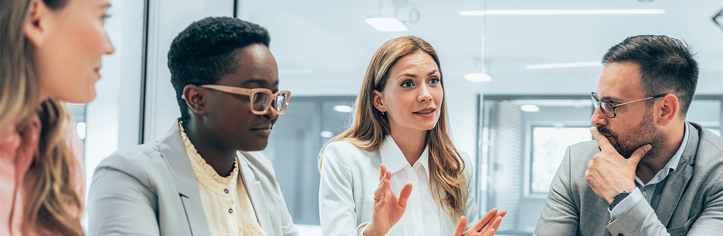 Four diverse professionals in business attire engaged in a meeting, with a woman in white speaking to attentive colleagues.