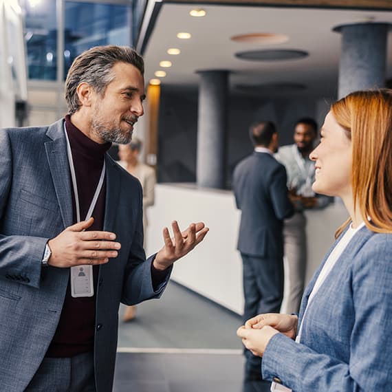Two business professionals having a conversation in a modern office lobby with colleagues in the background.