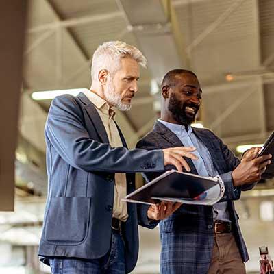 Two businessmen in suits reviewing documents together in a modern office, one pointing at a tablet while the other smiles.
