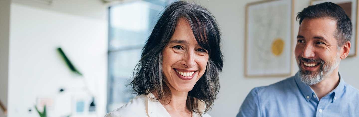 Smiling woman with dark hair in white shirt standing next to bearded man in blue shirt in bright office setting.
