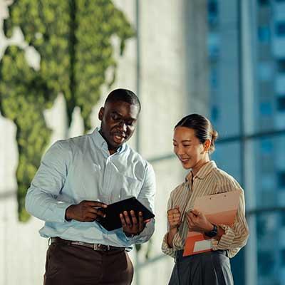 Two colleagues, standing in a modern office setting with a green foliage world map on the wall behind them. The man holds a tablet and gestures while speaking, and the woman smiles, holding a pink folder."