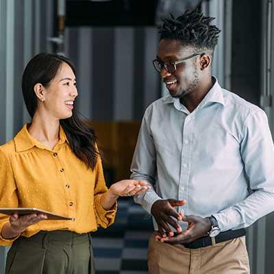 Two people walk together in a modern office hallway, engaged in conversation. One holds a tablet while gesturing with their other hand, and the other listens attentively with hands loosely clasped.